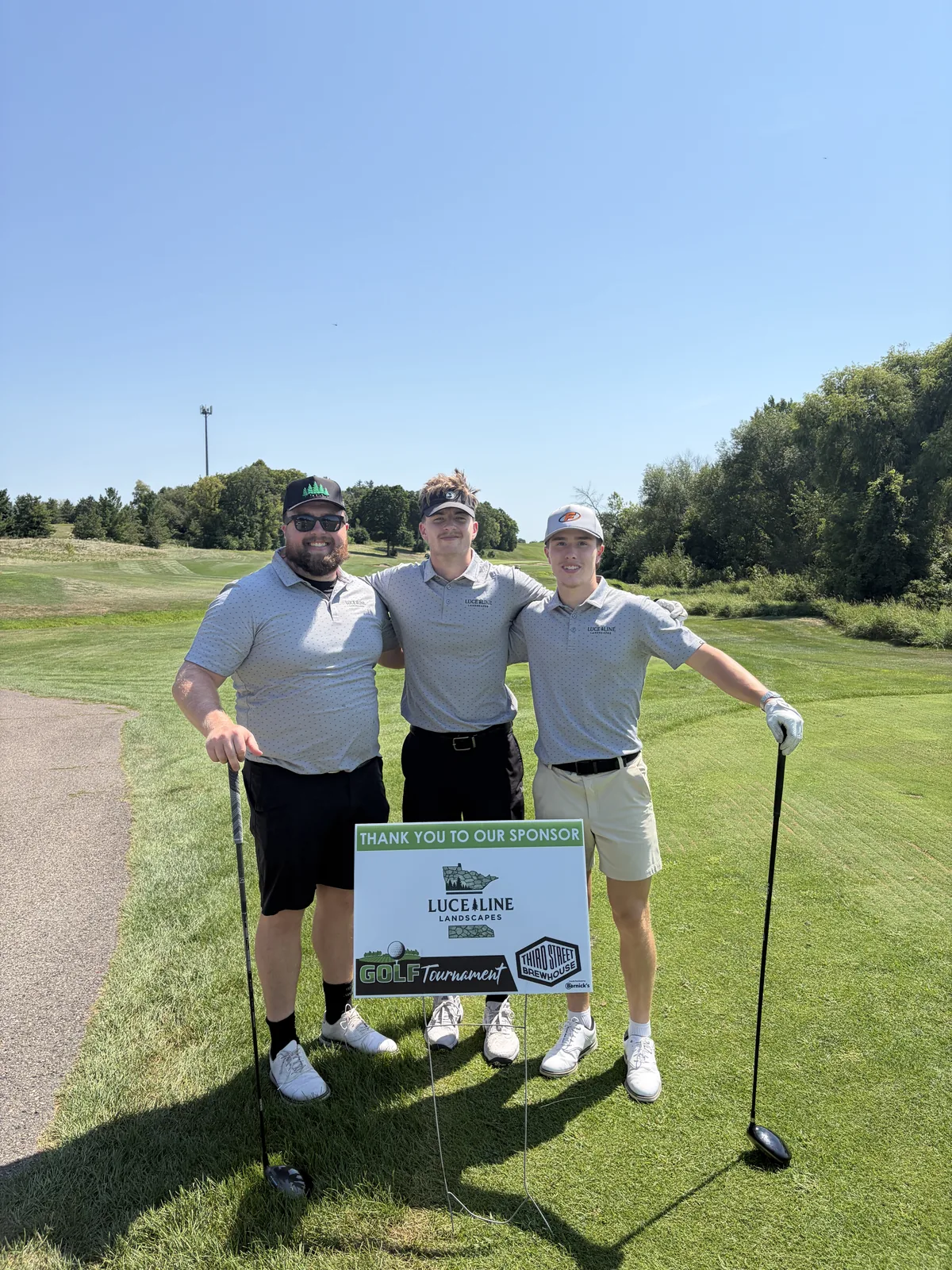 Three members of the Luce Line Landscapes crew at the Third Street Brewhouse golf tournament, beside a sign thanking Luce Line Landscapes as a sponsor.